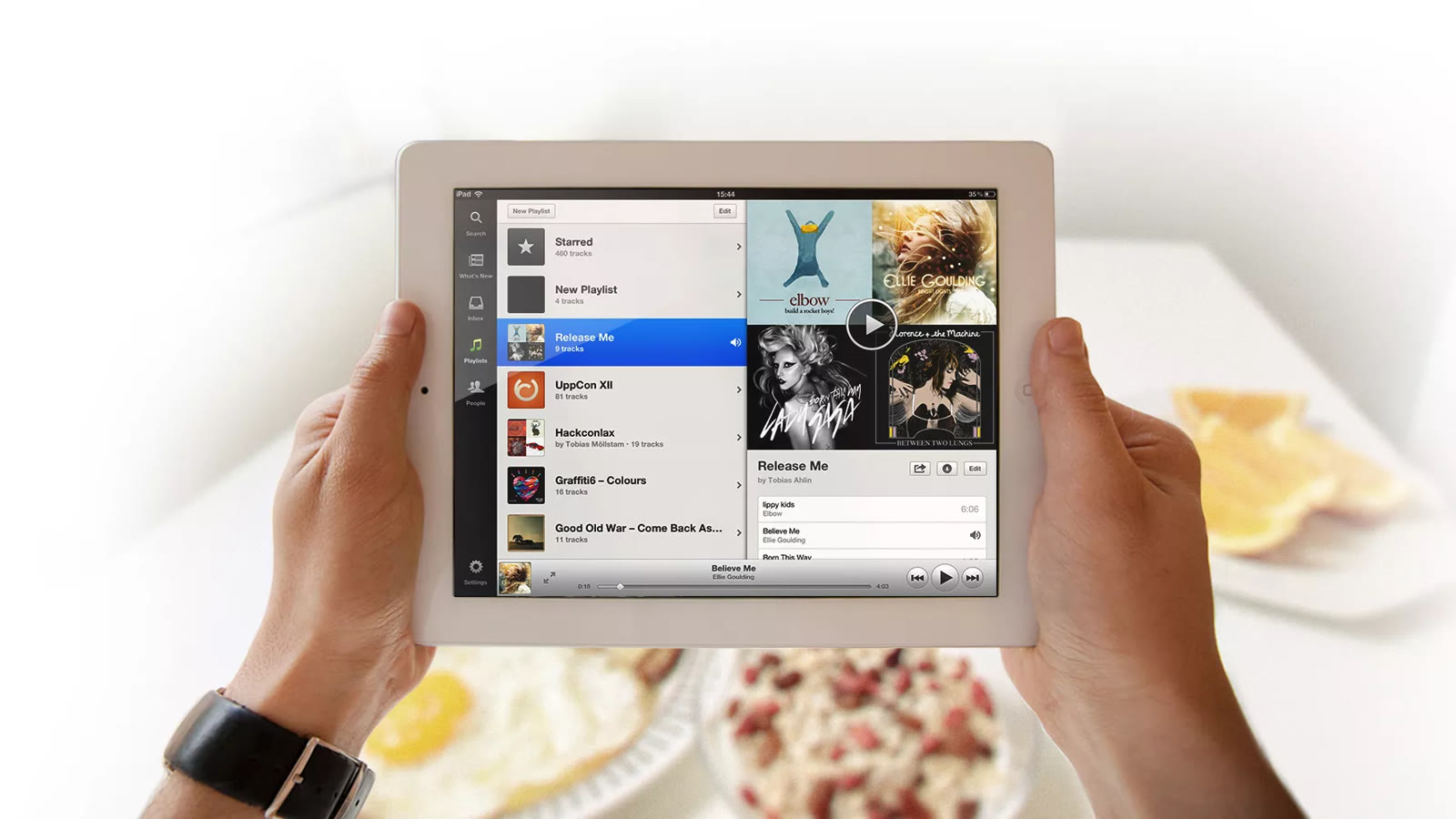 Two hands holding an iPad running Spotify, with the menu rail, a playlist and album covers on screen.